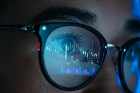Close up of a person wearing glasses. A blue computer screen with digital graphs in reflected in their glasses.